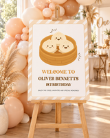 Little dumpling 1st birthday welcome sign on a wooden easel at the party entrance with a peach and cream balloon garland