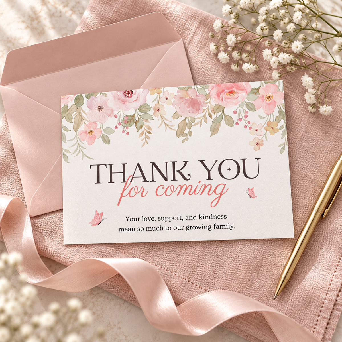 Flat lay of a pink floral baby shower thank you card with blush envelope and gold pen