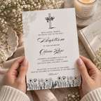 Black and white baptism invitation with an engraved cross and wildflower border, styled on a table