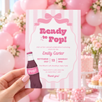 Hand holding the Ready To Pop baby shower invitation in front of pink balloons and a dessert table